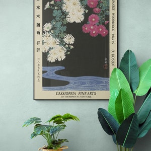 May include: A vintage Japanese woodblock print of white and pink chrysanthemums with a blue stream flowing through the bottom. The print is framed and has the text "JAPANESE WOODBLOCK PRINTS O. KOSON" and "CASSIOPEIA FINE ARTS 219 THOMPSON ST. NEW YORK" printed on it.