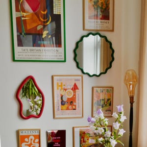 Op de afbeelding: Een galerijmuur met ingelijste kunstdrukken en decoratieve spiegels. De kunst omvat een Kandinsky-tentoonstellingsposter, een Monet-bloemenprint en een Matisse-print. Spiegels hebben rode en groene frames. Een vaas met bloemen staat op een tafel.