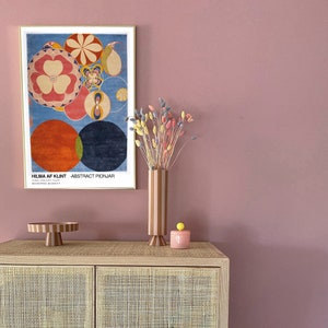 May include: A framed print of an abstract painting by Hilma af Klint titled "Abstract Pionjar". The painting features a colourful circular design with red, orange, yellow, blue, and green hues. A vase with dried flowers and a pink ceramic container with a yellow knob are on a wooden cabinet with a woven rattan front.