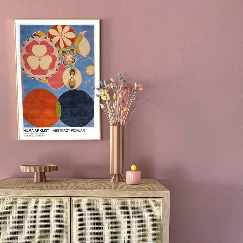 May include: A framed print of an abstract painting by Hilma af Klint titled "Abstract Pionjar". The painting features a colourful circular design with red, orange, yellow, blue, and green hues. A vase with dried flowers and a pink ceramic container with a yellow knob are on a wooden cabinet with a woven rattan front.