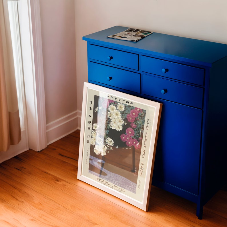 May include: A white framed print of a Japanese botanical illustration of white and pink flowers with the text "Japanese Woodblock Prints by A. Dawson" and "Cassiopeia Fine Arts". The print is leaning against a blue dresser with four drawers.