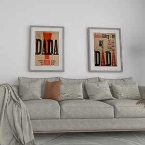 Op de afbeelding: Een grijze bank met een deken en kussens in een woonkamer. Twee ingelijste posters met het woord "DADA" in zwarte tekst op een witte achtergrond hangen aan de muur.