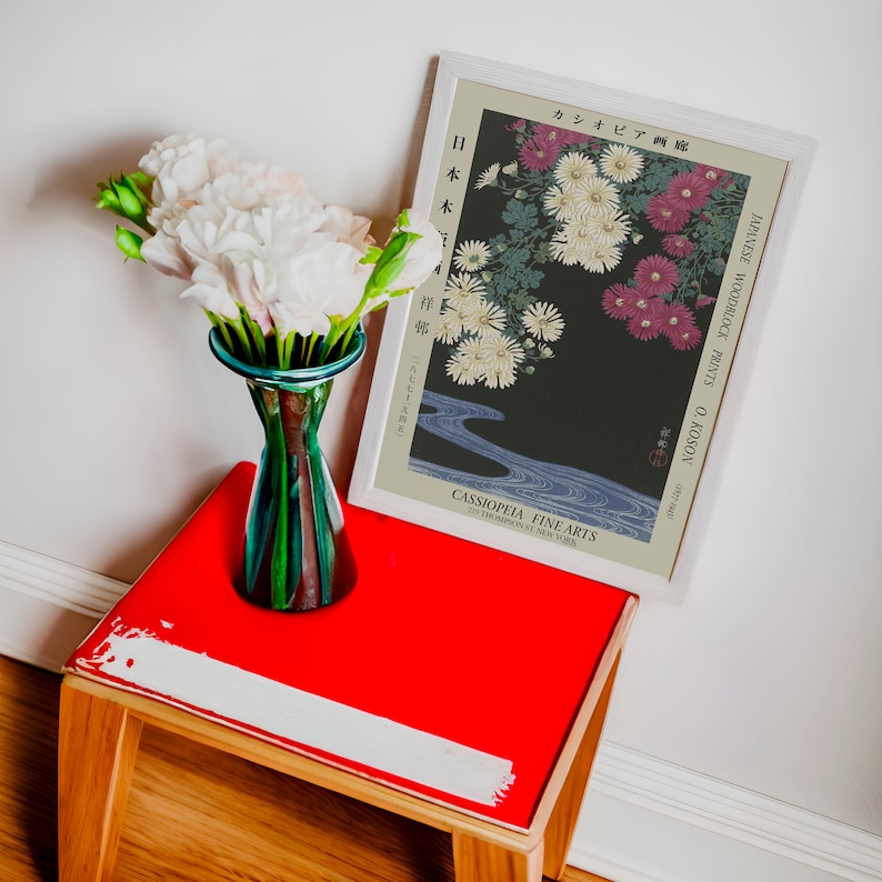 May include: A framed Japanese woodblock print of flowers and a river. The print is titled "Japanese Woodblock Prints O. Koson" and is from the gallery "Cassipoeia Fine Arts". The print is hanging on a white wall above a red table with a vase of white flowers.