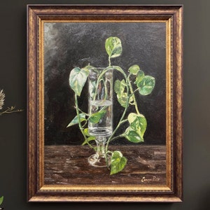 May include: A framed oil painting of a green plant growing in a clear glass vase. The vase is on a dark brown wooden surface. The painting is signed "Connie Riley" in the bottom right corner.