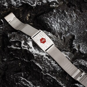 May include: A silver metal medical alert bracelet with a red medical symbol and a floral design. The bracelet is on a dark gray textured surface.