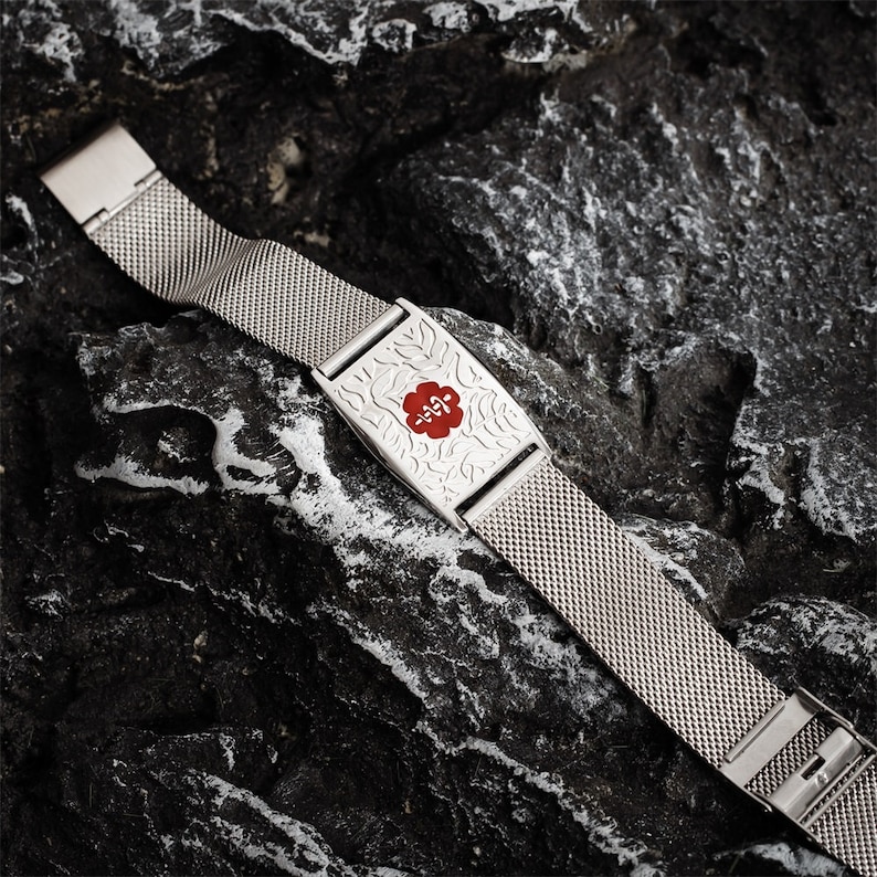 May include: A silver metal medical alert bracelet with a red medical symbol and a floral design. The bracelet is on a dark gray textured surface.