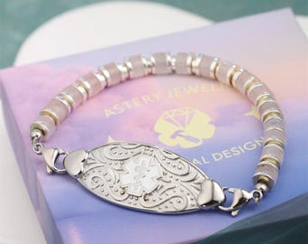 May include: A silver medical ID bracelet with a decorative oval plate featuring a white medical symbol. The bracelet has a beaded design with pink and gold-toned beads. The brand name "ASTERY JEWEL" is visible.