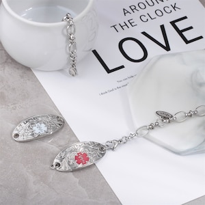 May include: A silver medical alert bracelet with two oval-shaped tags. One tag is white with a red medical symbol and the other tag is red with a white medical symbol. The bracelet has a lobster clasp and a small charm that says "LOVE".