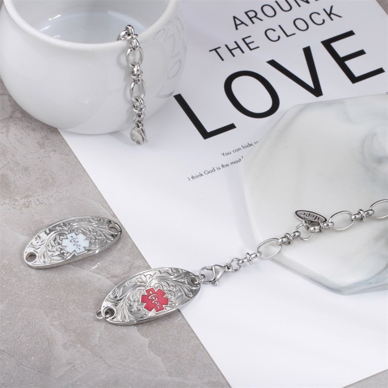 May include: A silver medical alert bracelet with two oval-shaped tags. One tag is white with a red medical symbol and the other tag is red with a white medical symbol. The bracelet has a lobster clasp and a small charm that says "LOVE".
