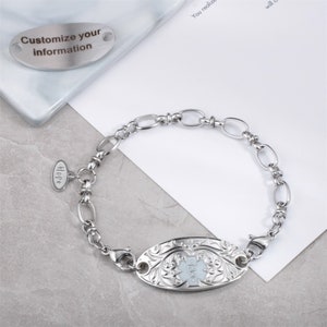 May include: A silver medical ID bracelet with a white enamel oval charm featuring a medical symbol and floral design. The bracelet has a small oval charm with the word "Hope" engraved on it. The bracelet is on a white surface.