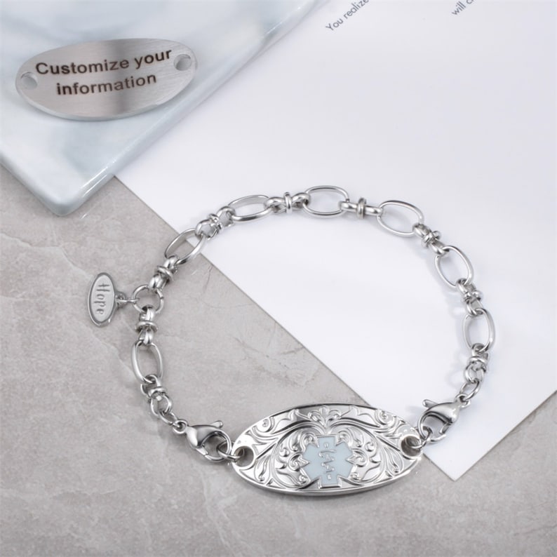 May include: A silver medical ID bracelet with a white enamel oval charm featuring a medical symbol and floral design. The bracelet has a small oval charm with the word "Hope" engraved on it. The bracelet is on a white surface.