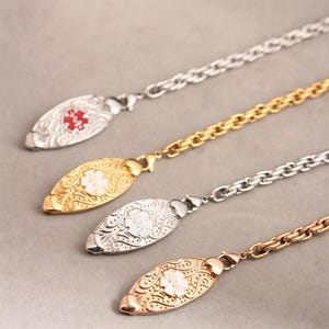 May include: Four medical alert necklaces featuring oval-shaped pendants with intricate scrollwork patterns. Each pendant displays a Star of Life emblem in red or white. The necklaces include silver-tone, gold-tone, and rose gold-tone metal chains.