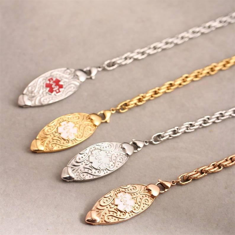 May include: Four medical alert necklaces featuring oval-shaped pendants with intricate scrollwork patterns. Each pendant displays a Star of Life emblem in red or white. The necklaces include silver-tone, gold-tone, and rose gold-tone metal chains.