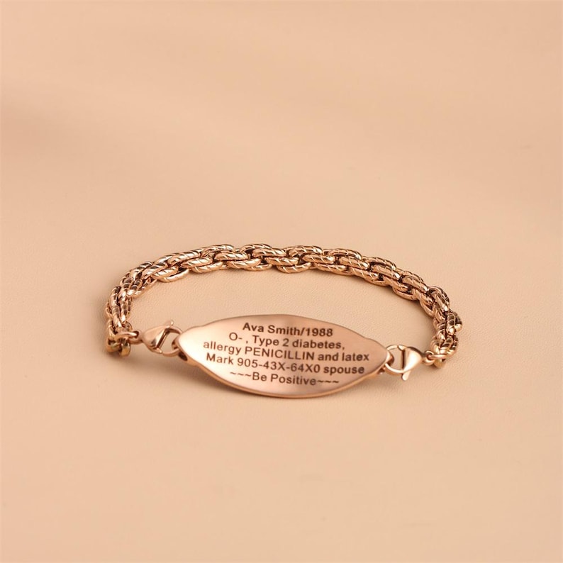 May include: A rose gold-toned medical alert bracelet featuring a textured rope-style chain and an oval-shaped identification plate. The plate is engraved with the text: Ava Smith/1988, O-, Type 2 diabetes, allergy PENICILLIN and latex, Mark 905-43X-64X0 spouse, ---Be Positive---.