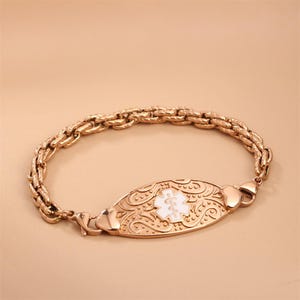May include: A rose gold-toned medical alert bracelet featuring a thick, textured chain and an oval-shaped central plaque. The plaque displays an embossed scrollwork pattern and a white enamel Star of Life symbol with a central staff and serpent.