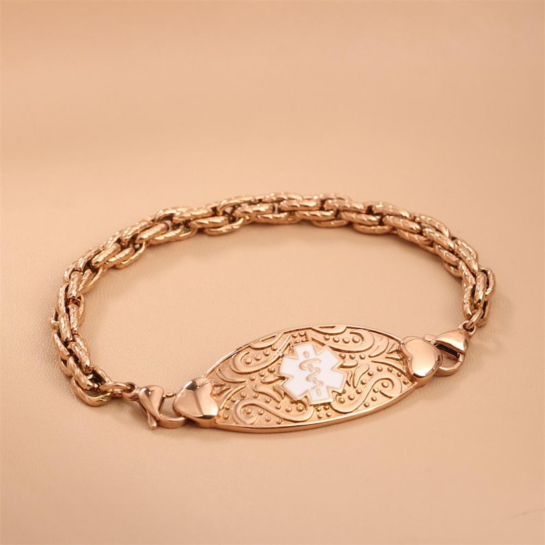 May include: A rose gold-toned medical alert bracelet featuring a thick, textured chain and an oval-shaped central plaque. The plaque displays an embossed scrollwork pattern and a white enamel Star of Life symbol with a central staff and serpent.