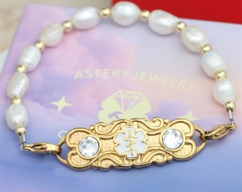 May include: A gold-toned medical ID bracelet with a white enamel medical symbol and two clear crystals. The bracelet features a strand of white, irregularly shaped pearls and gold-colored beads. The brand name "ASTERY JEWELRY" is visible.