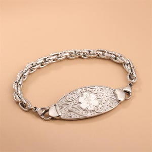 May include: A silver-toned medical alert bracelet featuring a thick, textured rope-style chain. The central oval plaque displays an embossed Star of Life symbol surrounded by intricate, swirling scrollwork patterns on a polished metallic surface.