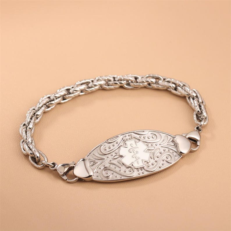 May include: A silver-toned medical alert bracelet featuring a thick, textured rope-style chain. The central oval plaque displays an embossed Star of Life symbol surrounded by intricate, swirling scrollwork patterns on a polished metallic surface.
