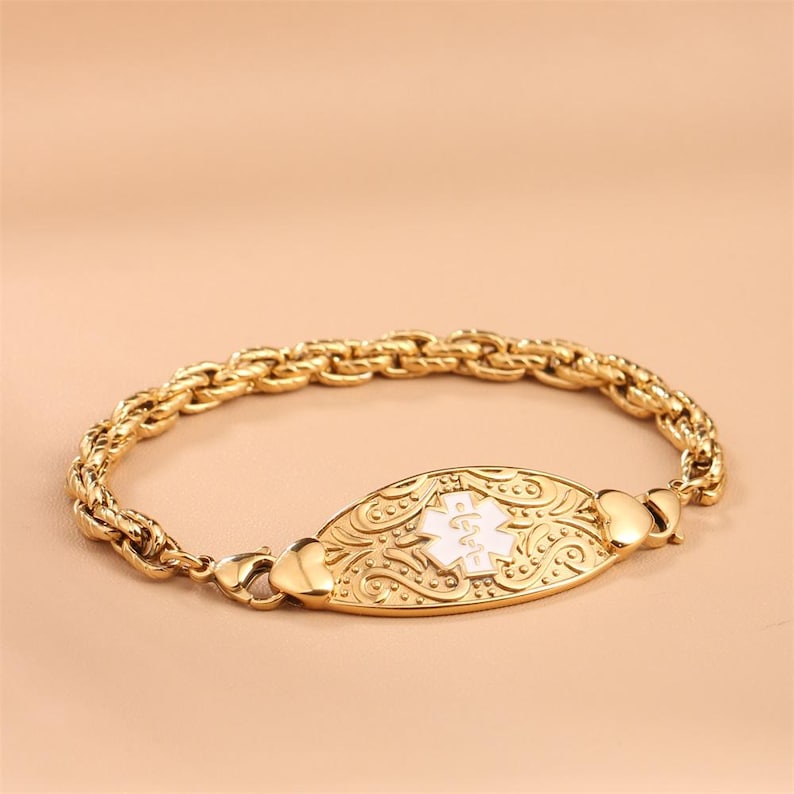 May include: A gold-toned medical alert bracelet featuring a twisted rope chain and a central oval plaque. The plaque displays an embossed scrollwork pattern and a white enamel Star of Life symbol with a serpent and staff emblem in the center.
