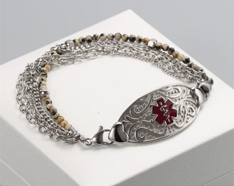 May include: A multi-strand medical alert bracelet featuring silver-toned metal chains and a string of small, speckled beige and black dalmatian jasper beads. The centerpiece is an oval silver-toned plaque with an embossed scroll pattern and a red medical star.
