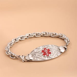 May include: A silver-toned medical alert bracelet featuring a thick, twisted rope-style chain. The central oval plaque displays an embossed scrollwork pattern and a prominent red Star of Life symbol with a white serpent and staff emblem.