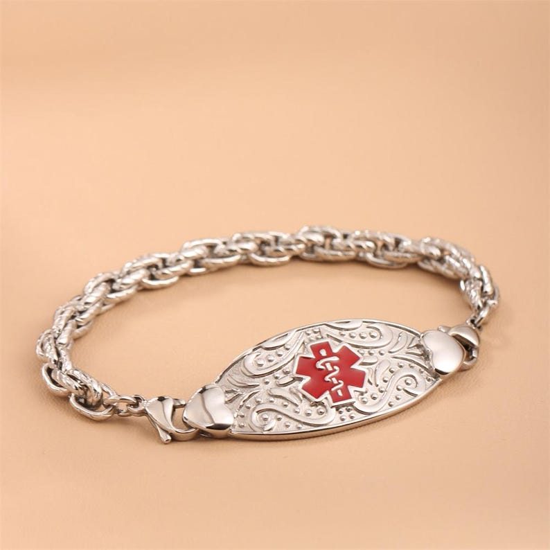 May include: A silver-toned medical alert bracelet featuring a thick, twisted rope-style chain. The central oval plaque displays an embossed scrollwork pattern and a prominent red Star of Life symbol with a white serpent and staff emblem.