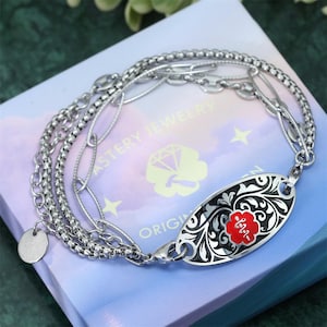 Könnte beinhalten: Ein silbernes Armband mit verschiedenen Kettenarten und einem ovalen Anhänger. Der Anhänger hat einen schwarzen Hintergrund mit floralem Muster und einem roten Blumenemblem. Das Armband liegt auf einer hellblauen Schachtel.