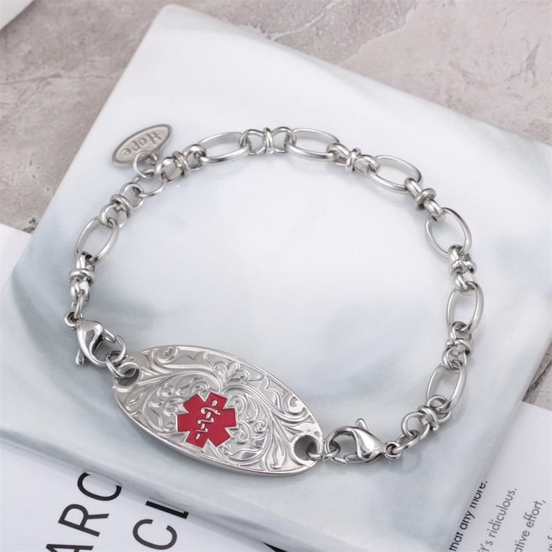 May include: Silver medical ID bracelet with a red medical symbol and a floral design. The bracelet has a lobster clasp and a small charm that says "Hope".
