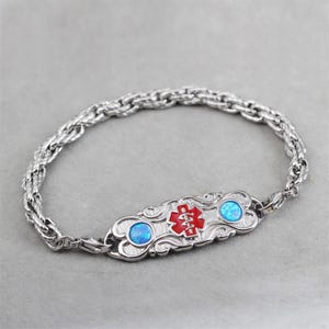 Puede incluir: Un brazalete de alerta médica de plata con un símbolo médico rojo y dos piedras de ópalo azules. El brazalete tiene un diseño de cadena de cuerda.