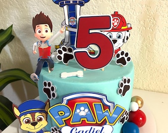 May include: A birthday cake decorated with Paw Patrol characters and a large number 5. The cake is light blue with paw prints and the Paw Patrol logo. Includes character cutouts of Chase, Marshall, and Ryder.
