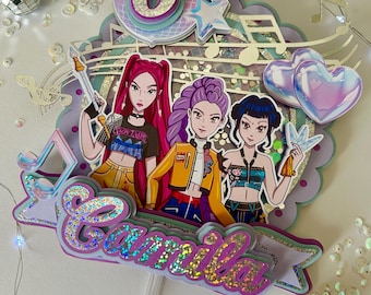 May include: A multi-layered cake topper featuring three cartoon figures with colorful hair and outfits. The topper includes a large number 6, stars, musical notes, and the name "Camila" in glittery letters. The overall design is vibrant and celebratory.