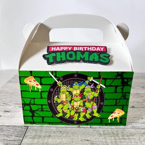 May include: A white cardboard party favor box with a green, black, and red Teenage Mutant Ninja Turtles design. The box has a handle and says "Happy Birthday Thomas" on the top.