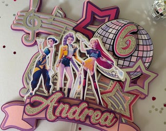 May include: A layered cake topper featuring three stylized figures, a musical note, stars, and a disco ball with the number 6. The name "Andrea" is displayed in glittery letters. The color scheme includes pink, purple, and silver.