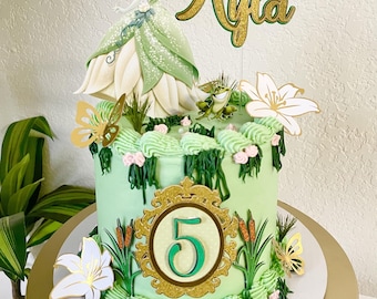 May include: A light green frosted cake decorated with a Princess Tiana topper, a gold glitter name topper reading Ayla, a firefly, a frog, white lilies, gold butterflies, and a central gold glitter frame containing the number 5.