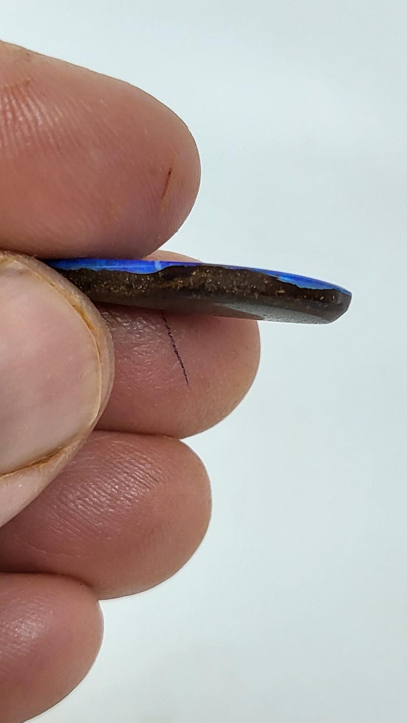 May include: A polished boulder opal gemstone featuring a vibrant, iridescent blue surface layer set against a dark, earthy brown ironstone matrix. The stone is flat and elongated, showcasing a distinct contrast between the bright blue play-of-color and the natural, textured dark brown base material.