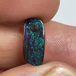 Puede incluir: Un cabujón de ópalo verde oscuro y negro con un alto pulido. El ópalo tiene una iridiscencia verde y azul.