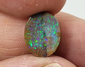 May include: An oval-shaped opal gemstone with a brown matrix and vibrant flashes of green, blue, and purple. The gemstone is held between fingers, showcasing its polished surface and natural color variations. A close-up view highlights the stone's unique patterns.