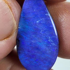 May include: A teardrop-shaped polished gemstone featuring a deep, vibrant blue base color with iridescent flashes of lighter blue and subtle white veining throughout the surface, displaying a smooth, reflective finish across the entire stone.