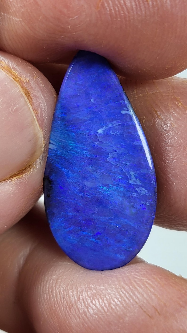 May include: A teardrop-shaped polished gemstone featuring a deep, vibrant blue base color with iridescent flashes of lighter blue and subtle white veining throughout the surface, displaying a smooth, reflective finish across the entire stone.