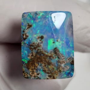 May include: A rectangular polished opal gemstone featuring a vibrant play of color with iridescent flashes of blue and green against a dark base. The stone displays natural brown matrix inclusions throughout its surface, creating a contrast.