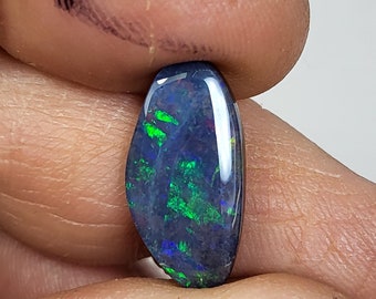 May include: A polished, oval-shaped black opal gemstone. The opal displays vibrant flashes of green and blue within its dark surface. The gemstone is held between two fingers against a white background.