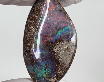 May include: A polished, teardrop-shaped boulder opal gemstone featuring a dark brown ironstone matrix with vibrant flashes of red, green, and blue iridescent color play across its smooth, reflective surface, showcasing natural geological patterns.