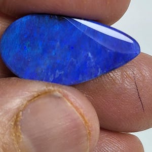 May include: A pear-shaped, polished gemstone featuring a deep, vibrant blue color with subtle internal flashes of lighter blue and violet. The stone has a smooth, reflective surface and a distinct teardrop silhouette, showcasing a rich, saturated hue.