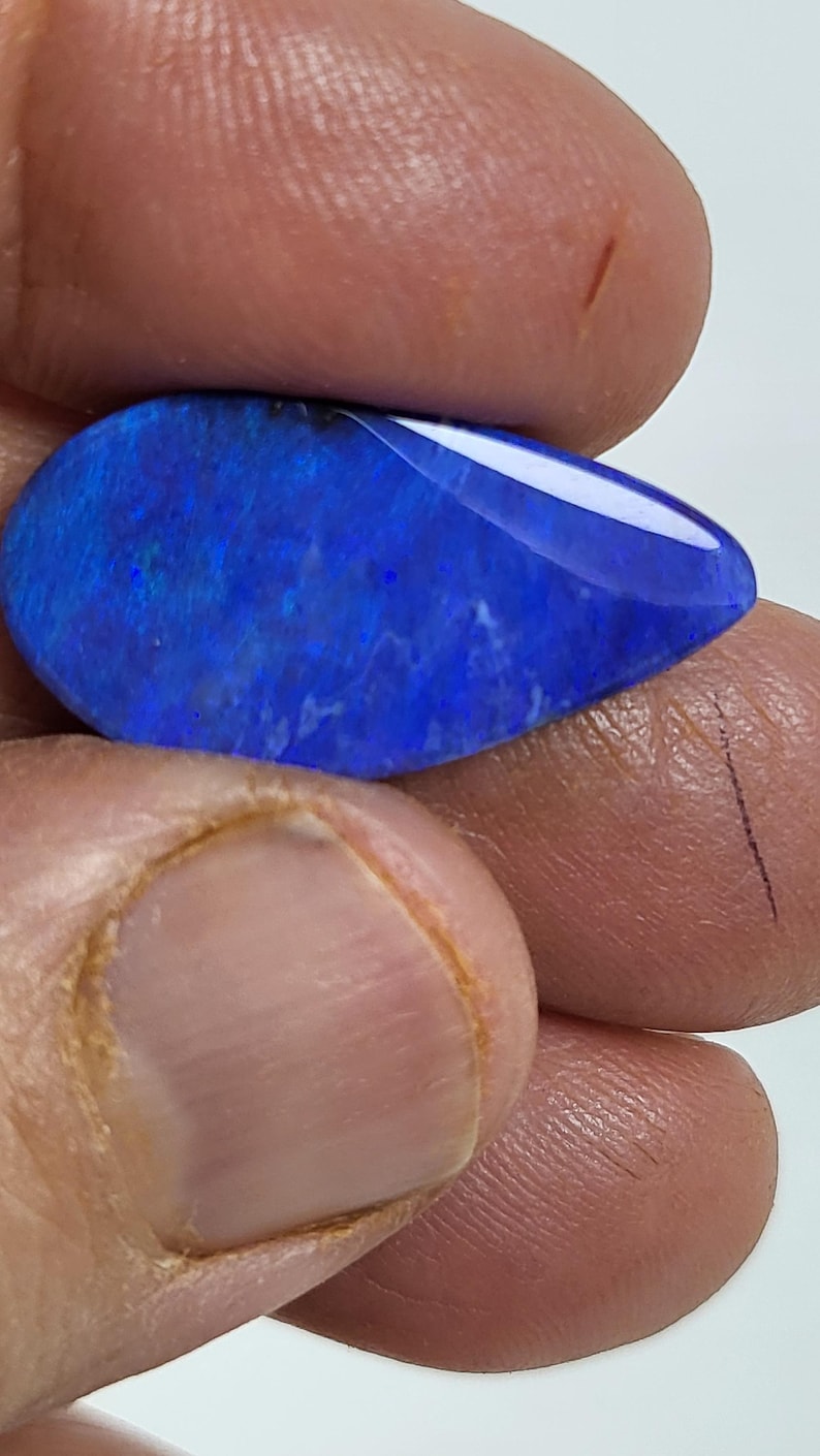 May include: A pear-shaped, polished gemstone featuring a deep, vibrant blue color with subtle internal flashes of lighter blue and violet. The stone has a smooth, reflective surface and a distinct teardrop silhouette, showcasing a rich, saturated hue.
