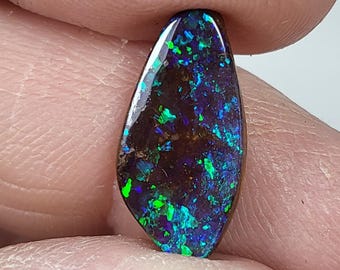 May include: A polished, teardrop-shaped opal gemstone. The stone displays a dark base color with vibrant flashes of blue, green, and purple. The opal is held between two fingers, showcasing its iridescent play-of-color.