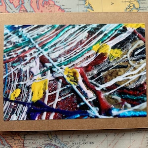 May include: Abstract art print featuring a close-up of colorful paint drips and splatters. The artwork showcases a mix of red, white, yellow, blue, and silver metallic paint on a textured surface, presented within a brown card.