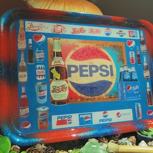 May include: A rectangular rolling tray with a glossy finish, featuring a red and blue gradient border. The centre displays various vintage Pepsi-Cola logos, imagery of glass bottles, aluminium cans, and a six-pack carton, all centred around the Pepsi logo.
