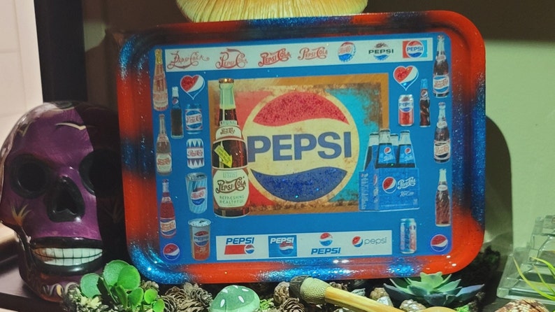 May include: A rectangular rolling tray featuring a collage of vintage Pepsi branding, including various soda bottles, cans, and the classic Pepsi logo. The tray has a glossy finish with a red and blue gradient border and rounded corners.