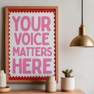 May include: A framed print with the words "YOUR VOICE MATTERS HERE" in pink block letters. The print has a white background with a scalloped edge and a red border, set in a wooden frame. A gold-colored pendant light hangs above.